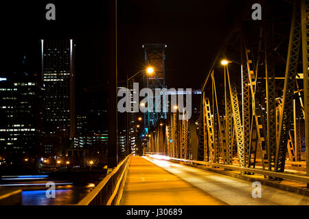 Evening lights lit metal drawbridge with two supporting towers across the river on a background of a passing downtown city blurred silhouette Stock Photo