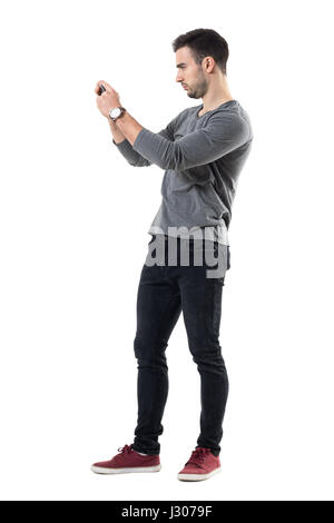 Profile view of serious young casual man holding cellphone taking photo. Full body length portrait isolated over white studio background. Stock Photo