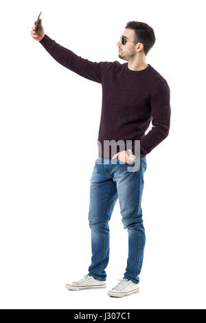 Side view of handsome casual man with sunglasses taking selfie with cell phone. Full body length portrait isolated over white studio background. Stock Photo