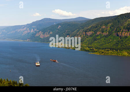 Two barges float towards each other on a wide Columbia River in Washington state, surrounded by picturesque mountains covered with green trees. Stock Photo