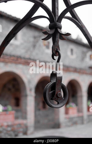 Old rusty metal block suspended from the arched wrought-iron grille over the well in an old castle on a background of the arched gallery of stone Stock Photo