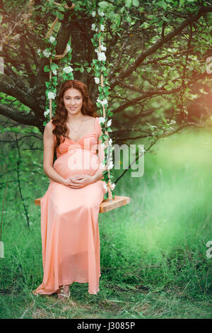 Young beautiful pregnant girl with long brown hair in peach dress with flower wreath on her head in swing near tree. Stock Photo