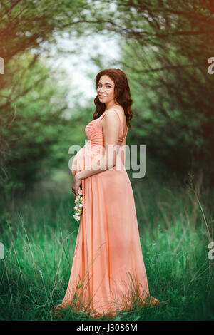 Young beautiful pregnant girl with long brown hair in salmon dress in green garden Stock Photo