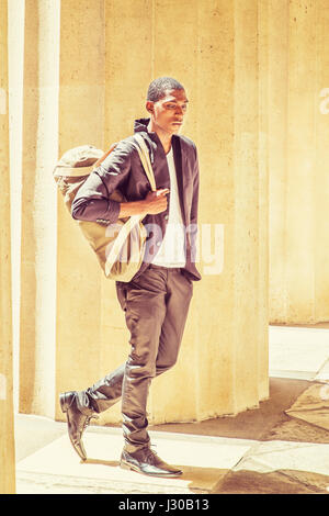 Young African American Man traveling in New York, carrying shoulder bag, walking though columns on campus, looking down, thinking. Street Fashion. Ins Stock Photo