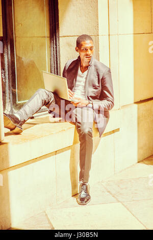 Young African American Man traveling, studying in New York, wearing black fashionable jacket, white v neck undershirt, pants, leather shoes, sitting a Stock Photo