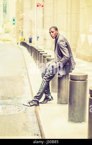 Young black teenage boy is sitting on street, hunchbacked, sad, tired, looking down, thinking, lost in thought. Man relaxing on street. Retro filtered Stock Photo