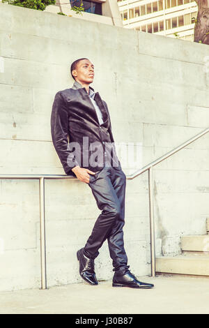 City Life. Wearing a fashionable jacket, pants, leather shoes, a young black college student standing against the wall, sad, thinking. Concept of teen Stock Photo