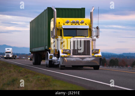 Classic American powerful yellow big rig semi truck with high chrome tailpipes powerful headlights and green container local cargo driving on road Stock Photo