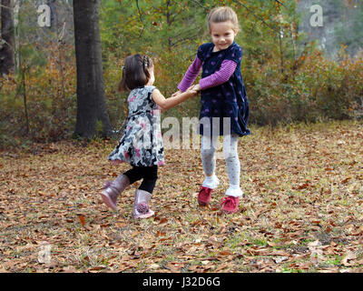 Two little girls hold hands and swing in circles.  They are playing outdoor games in the Fall leaves. Stock Photo