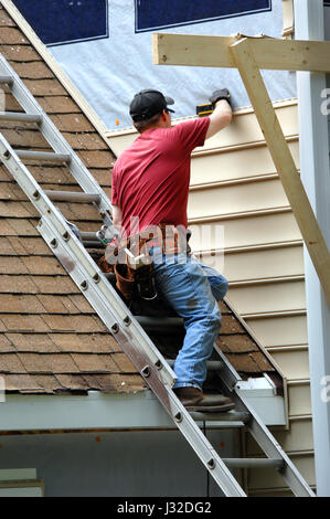 Young carpenter remodels aging home.  He is installing siding and is using a level to be sure installation is precise.  He is standing on a ladder on  Stock Photo