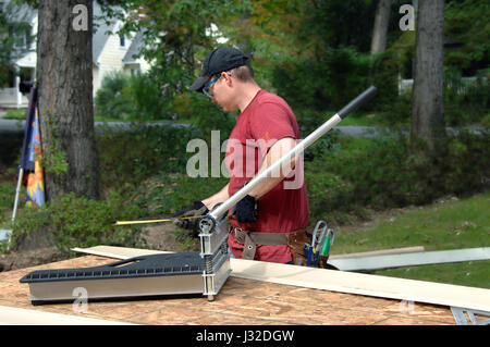Young man works to put siding on his home.  He is using a tape measure to check accuracy of his cut.  His is wearing a red T-shirt, safety glasses and Stock Photo