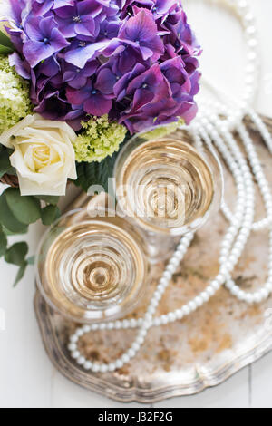 Big bouquet of fresh flowers, purple hydrangeas and white roses in a wicker basket, two glasses of champagne and vintage wedding decor with pearls on  Stock Photo