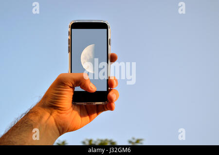 Man's hand taking a picture of the moon on a blue sky during the day with a smartphone Stock Photo
