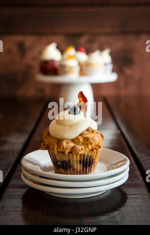 Close-up of a bacon blueberry breakfast cupcake on a stack of white plates, with a display of gourmet cupcakes in the background Stock Photo