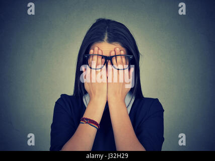 Closeup portrait young woman in glasses covering face eyes using her both hands isolated on gray wall background Stock Photo