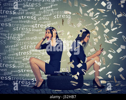 Two women sitting on a tire one being stressed another successful using mobile phone under money rain Stock Photo