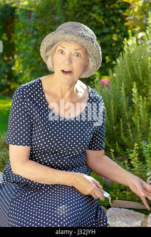 Senior white woman in a blue polka dotted and straw hat in her garden yard showing an exaggerated surprise, joy in garden concept Stock Photo