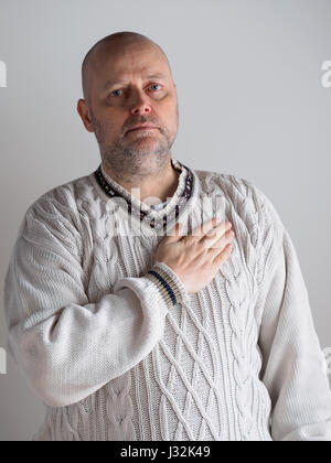 Portrait, casual looking caucasian male wearing a white pullover, right hand over heart and he looks toward the camera Stock Photo