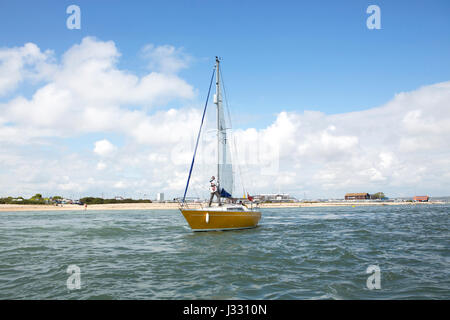Small yacht leaving Langstone harbour. Person standing on deck trimming the sails. Stock Photo