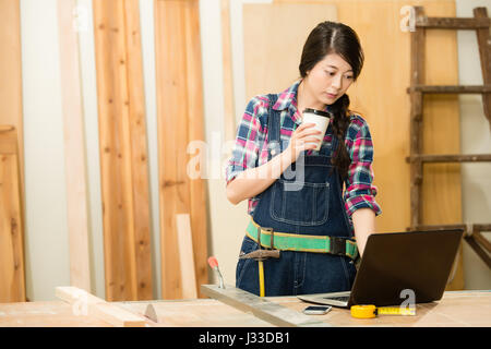 beautiful woman craftsman who owns a small business, bending over at work bench to type on laptop drinking coffee, while working in her workshop and d Stock Photo