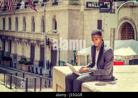 Young African American man traveling, working in New York, sitting on street by vintage office building, reading, working on laptop computer. Wall Str Stock Photo