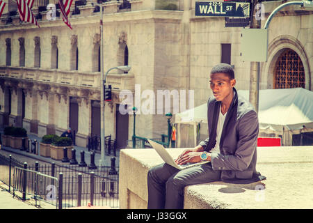 Young African American man traveling, working in New York, sitting on street by vintage office building, reading, working on laptop computer. Wall Str Stock Photo
