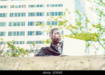 Young African American college student standing by top of  wall outside office building on campus in New York in spring day,  working on laptop comput Stock Photo