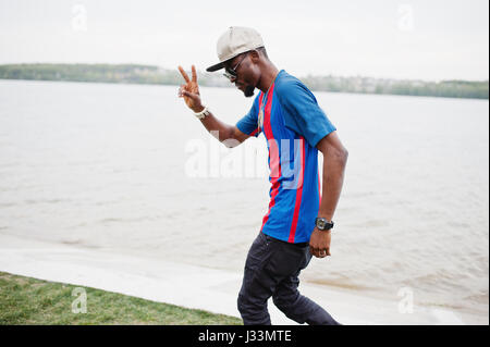 Stylish african american boy wear at cap, football t-shirt and sunglasses showing two fingers. Black sports man portrait. Stock Photo