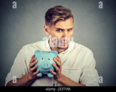 Young greedy stingy business man holding piggy bank isolated on gray wall background Stock Photo