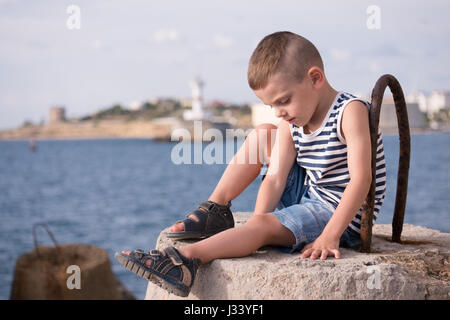 adorable little boy in vest and shorts sitting against the sea in summer Stock Photo