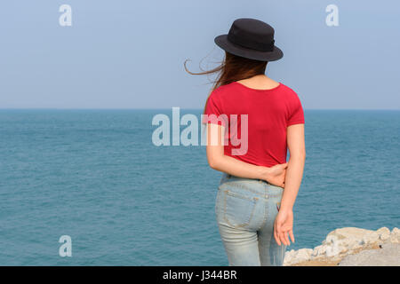 woman standing behind wearing red t-shirt and jeans and black hat with sea and blue sky background Stock Photo