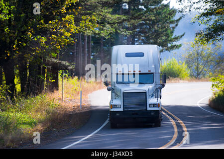 White classic powerful big rig long haul semi truck with a high roof transporting cargo on a bend of the winding road passing through the forest Stock Photo