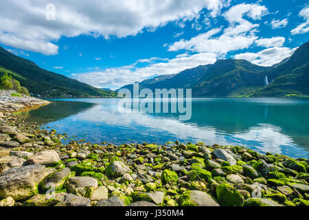 Rocky beach, moss covered rocks, blue sky, blue waters and a tall waterfall in the background Stock Photo