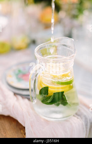 picnic, food, summer, holiday concept - adding of pure water for fresh lemonade, slices of lemon, lime, green mint petals in a glass jug on the table, fruit and plate on a background, selective focus. Stock Photo