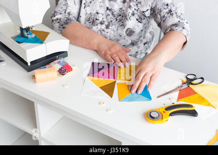 needlework and quilting in the workshop of a woman tailor - hands of tailor women put on the desktop the pieces of colored fabric, lay next to the scissors, buttons, pins and threads. Stock Photo