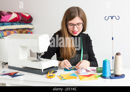 needlework and quilting in the workshop of a young woman, a tailor - young woman in glasses tailor at work with pieces of colored cloth on the table with threads, fabrics, needles and sewing machine Stock Photo