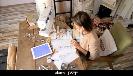 From above shot of young woman using tablet while sitting at table and drawing sketch in parlour. Stock Photo