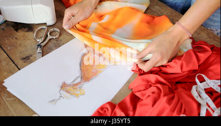 Crop female using sketch with drawn dress and choosing vibrant fabric in atelier. Stock Photo