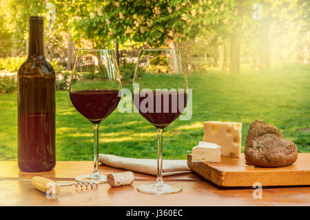 Glasses of red wine and a bottle, with slices of cheese at a picnic, on a wooden table, with green trees and grass in the blurred background, with a r Stock Photo