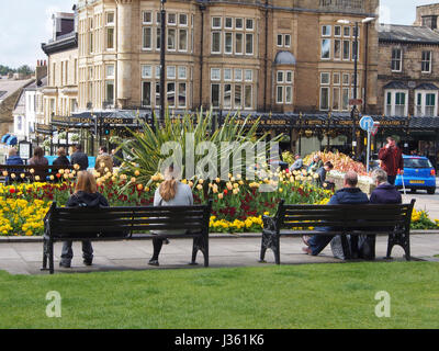 People relaxing in the sun in gardens in the centre of the Montpellier District of Harrogate, Yorkshire, UK, with Betty's Tea Rooms in the background. Stock Photo