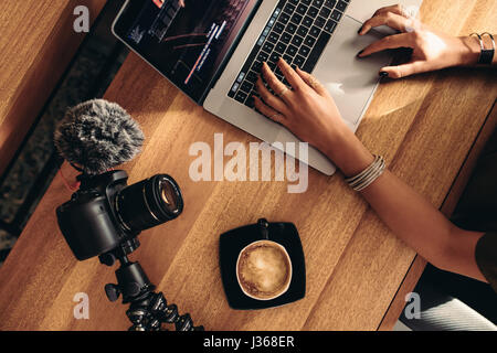 Top view of female vlogger editing video on laptop. Young woman working on computer with coffee and camera on table.