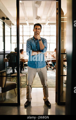 Full length portrait of creative young man standing in doorway of office. Caucasian male executive in office with colleagues working in background. Stock Photo