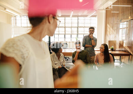 Business team sitting in conference room with female executive standing in front of glass wall with sticky notes. Stock Photo