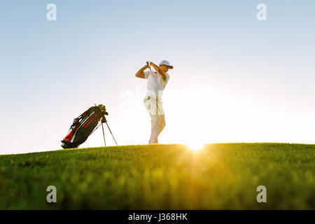 Low angle shot of male golfer taking shot while standing on field. Full length of golf player swinging golf club on sunny day. Stock Photo