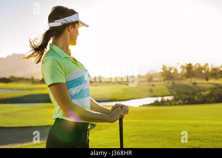 Professional female golfer holding golf club on field and looking away. Young woman standing on golf course on a sunny day. Stock Photo