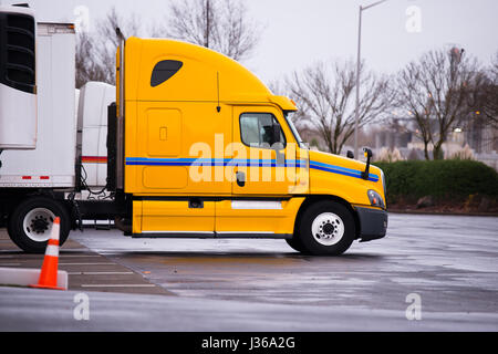 Profile of yellow modern professional big rig semi truck with a reefer trailer with a refrigeration unit is parked near a warehouse in anticipation Stock Photo