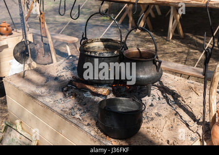 Black cauldrons hang on hooks over the open fire in a medieval camp. Cooking dinner for noble knights. Nobody around. Stock Photo