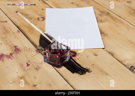 Inkpot with red ink inside, goose quill and a sheet of paper on a sunlit wooden table. Nobody around. Stock Photo