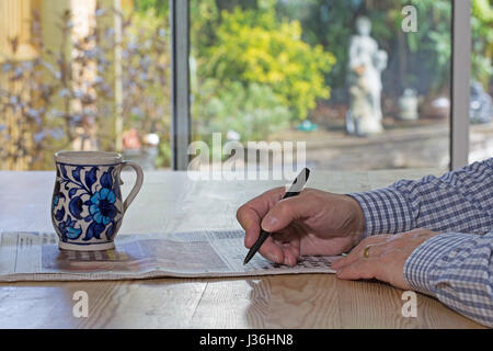 arms and hands of a man holding a pen whilst completing a newspaper crossword with coffee mug on the table and soft focus outdoor scene behind. Stock Photo