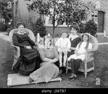 Portrait of the females from an Edwardian family, three adults and two children, sitting in a garden in 1910. On the left an older lady sits in a wicker armchair, sitting on the floor in front of her is a younger lady, possibly her daughter. Sitting in a wicker sofa are two young girls, one of which has a toddler, who is holding a trumpet, on her knee.Restored using a high resolution scan taken from the original negative. Stock Photo